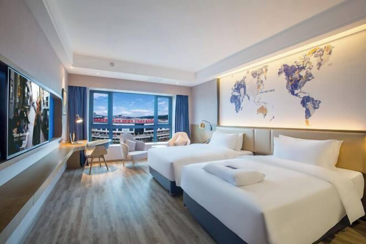A hotel room with two beds, a desk, a chair, a large window with a view, and a world map on the wall.