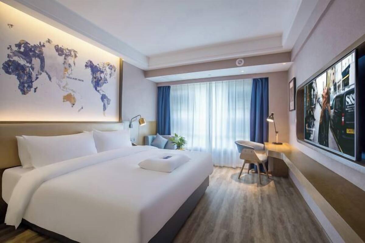 A hotel room with a large bed, a desk, a chair, a world map on the wall, and a large window with curtains.