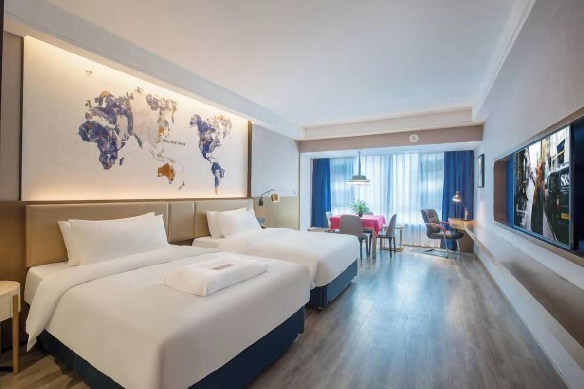 A hotel room with two beds, a world map on the wall, a dining area with a table and chairs, and a large window with curtains.