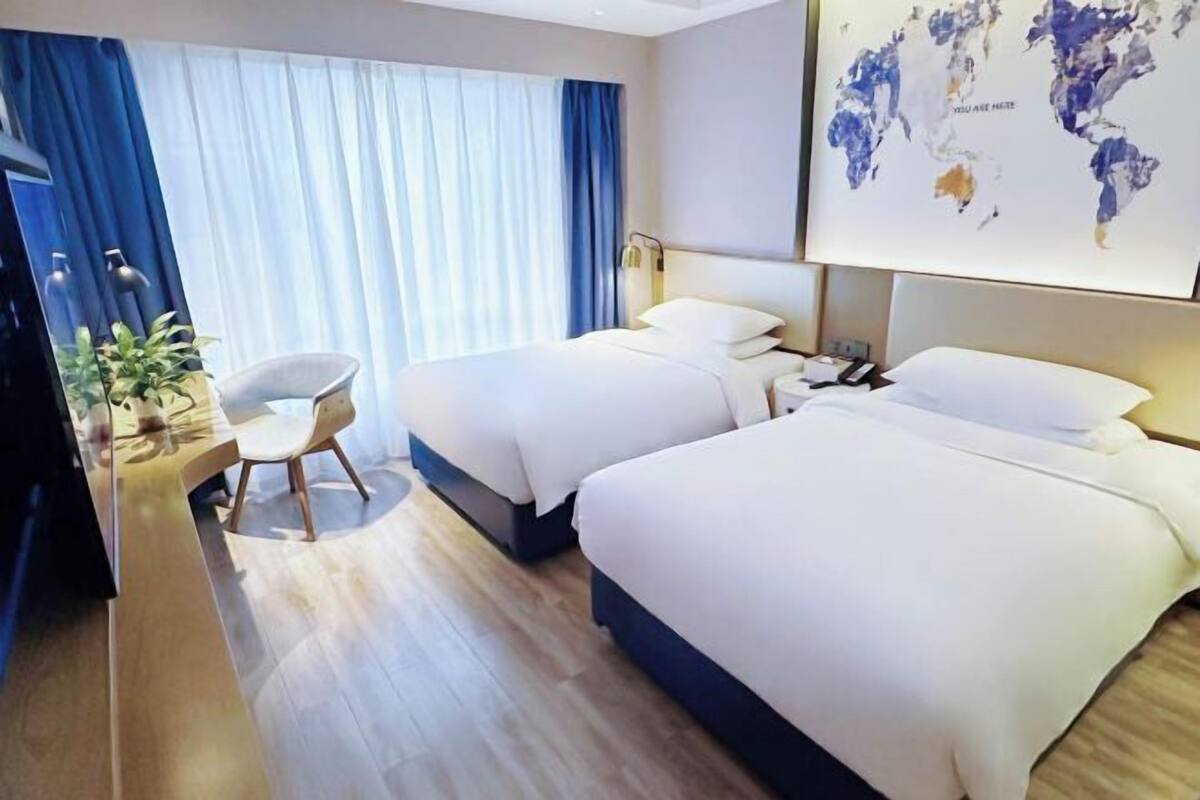 A hotel room with two beds, a desk with a chair, a world map on the wall, and a large window with curtains.