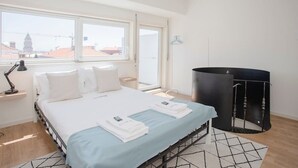 A modern hotel room with a large bed, a black radiator, a window with a view of buildings, and a small desk with a lamp. - Liiiving in Porto - Downtown Balcony Apartment (Porto)