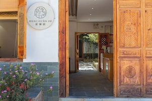 Una entrada de madera con un letrero, un camino que conduce al interior y una planta en maceta con flores rosadas.