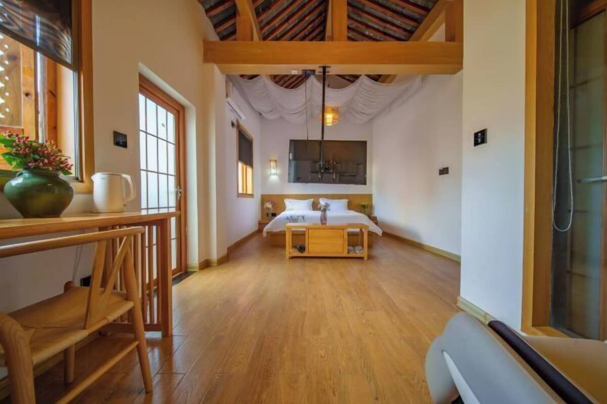 A bedroom with a wooden bed, a bedside table with a vase, a window, and a wooden ceiling.