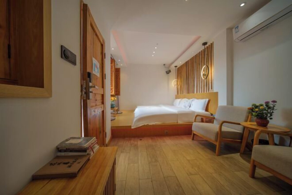 A modern hotel room with a wooden floor, a bed with white linens, a wooden headboard, a seating area with a chair and a small table, and a potted plant.