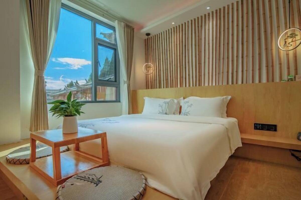 A modern hotel room with a large bed, a wooden headboard, a bedside table with a plant, and a view of a mountain landscape through a large window.