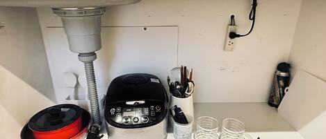 A kitchen shelf with a rice cooker, a red pot, a set of chopsticks, a can of spray paint, and a pair of scissors.