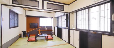 A traditional Japanese room with tatami flooring, a low table with cushions, sliding doors, and a TV on the wall.
