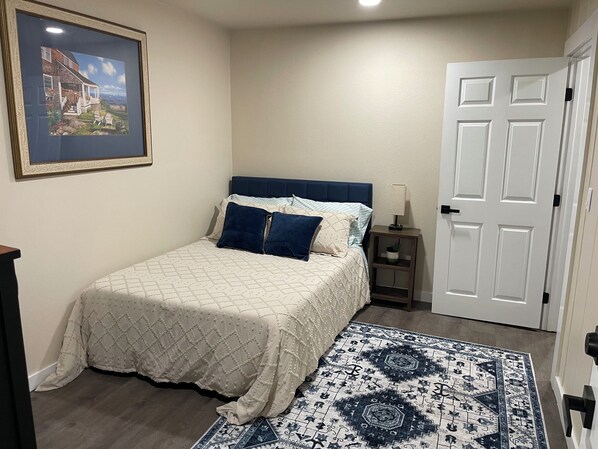 Un dormitorio con cama, mesita de noche, una puerta y un cuadro en la pared.