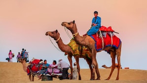 A camel caravan with riders in traditional attire, traversing a sandy terrain. - Ozaki Desert Camp Jaisalmer (Jaisalmer)