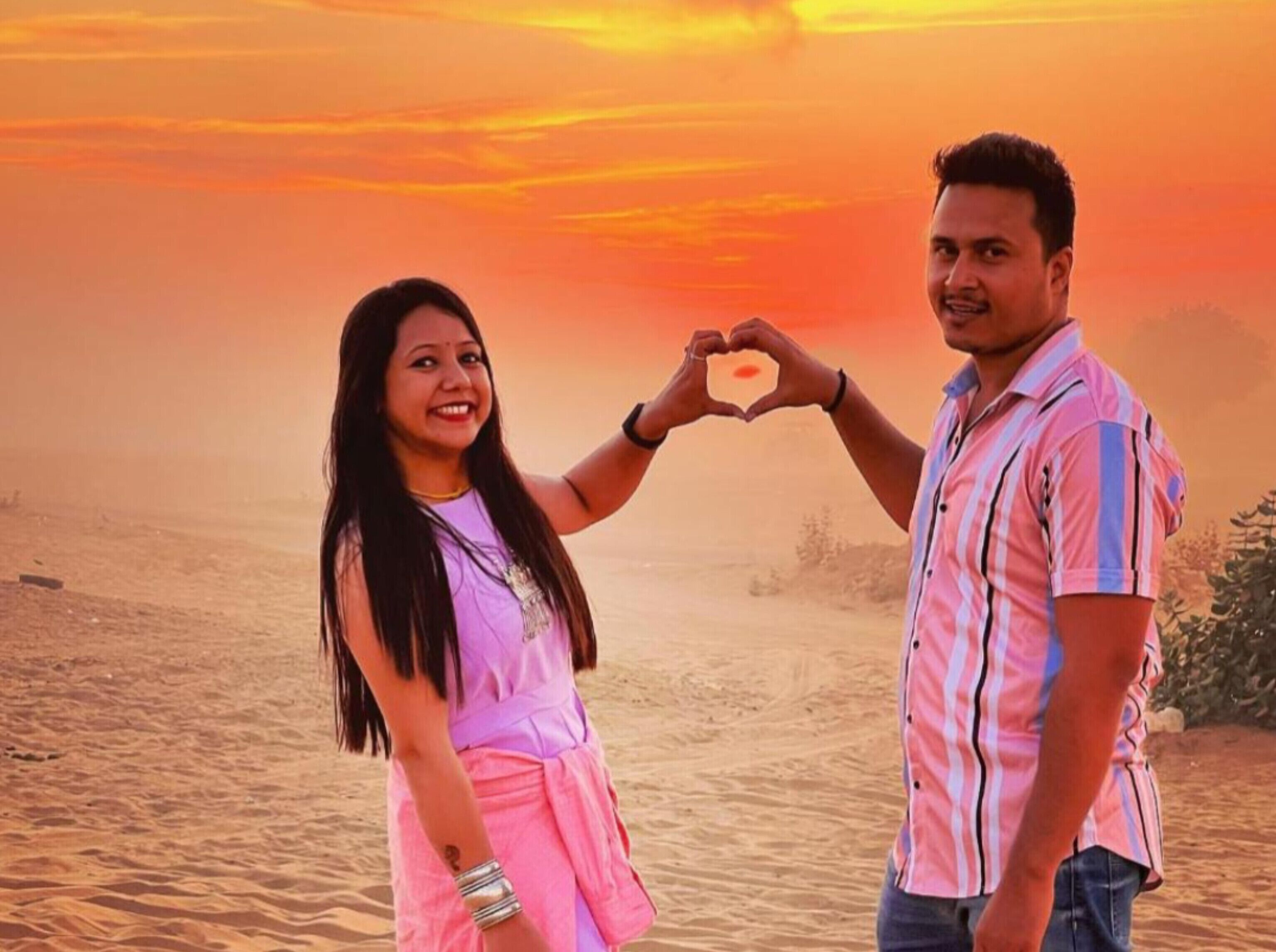 Un couple forme un cœur avec ses mains, le tout se détachant sur le fond d’un coucher de soleil.