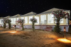 Tents with large windows and potted plants line the sandy ground, each lit by a lantern.