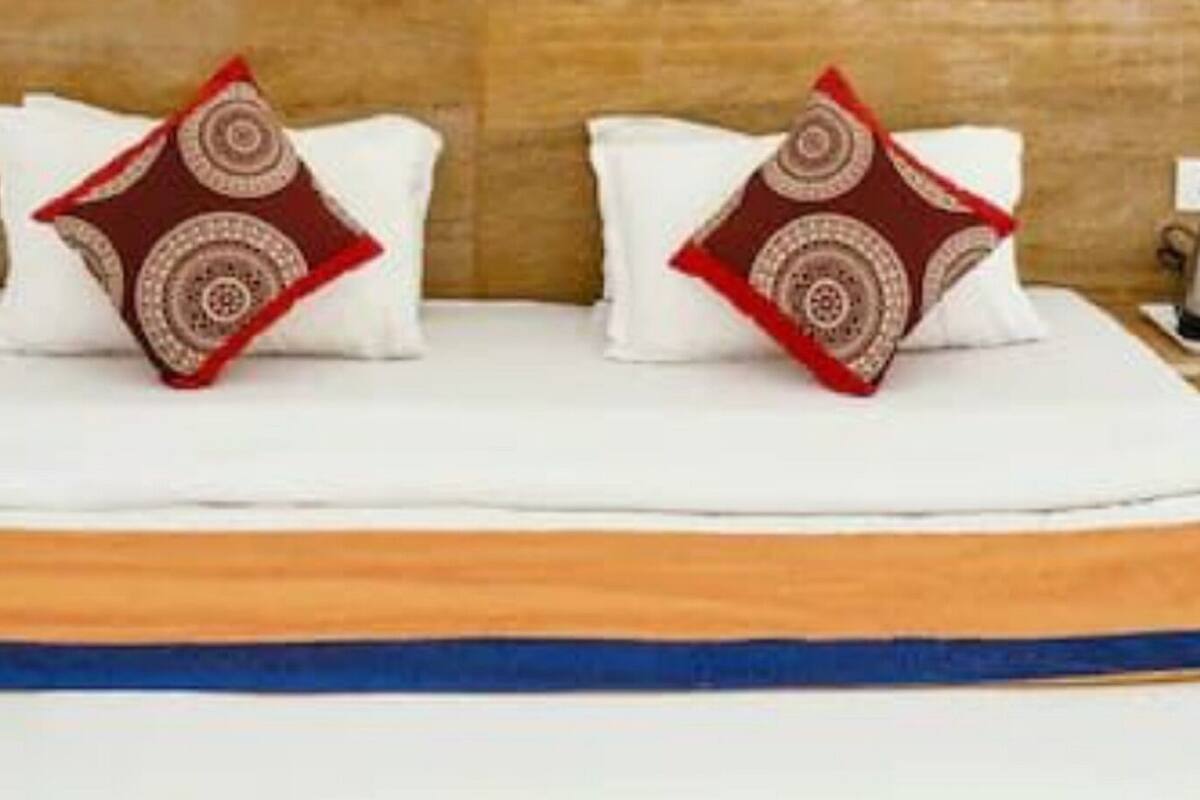 A hotel room with a bed, two decorative pillows, a bedside table with a kettle, and a wall with a patterned design.