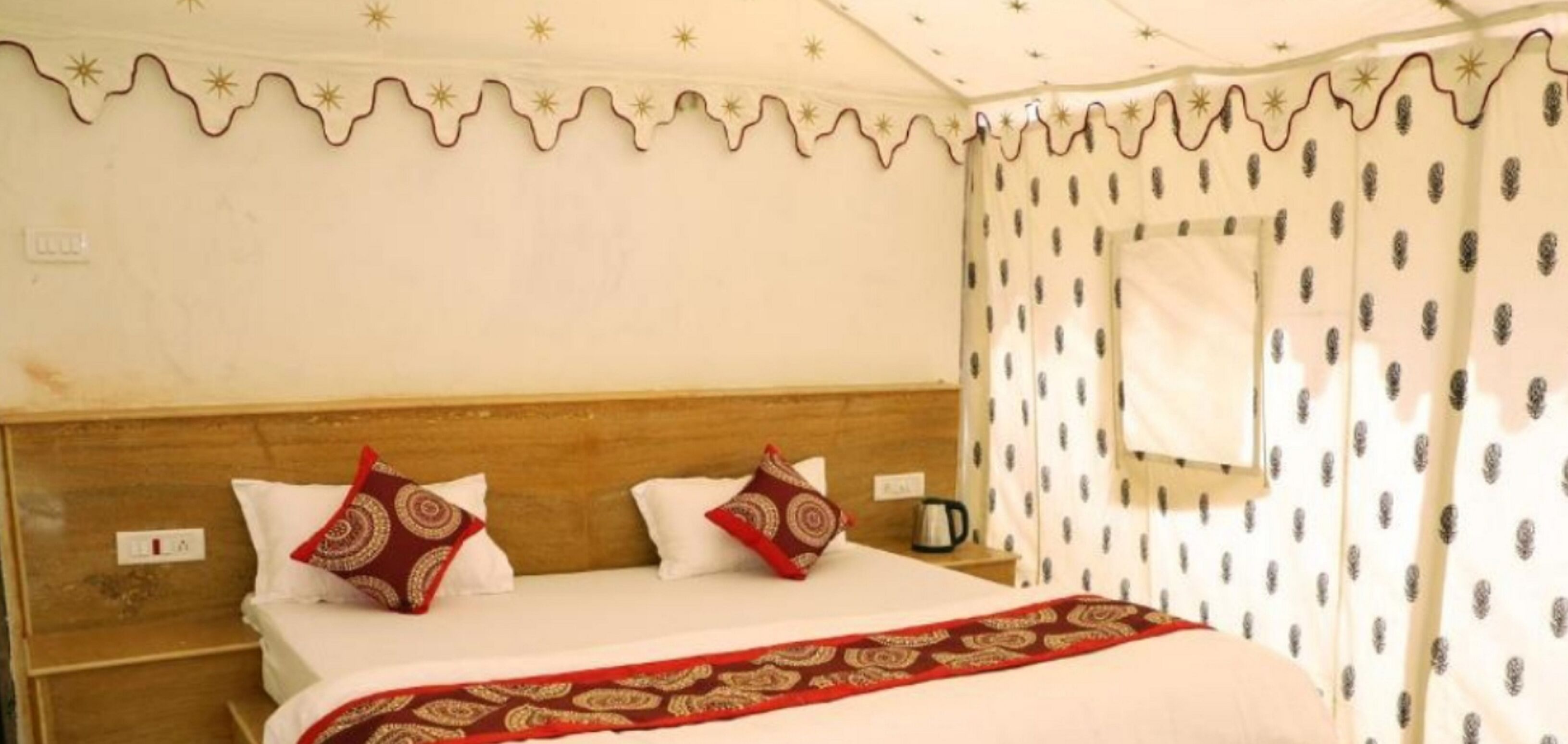 A hotel room with a wooden headboard, two white pillows, a red patterned throw blanket, and a nightstand with a coffee pot.
