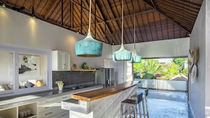 A modern kitchen with a wooden island, hanging teal pendant lights, and a view of greenery outside.
