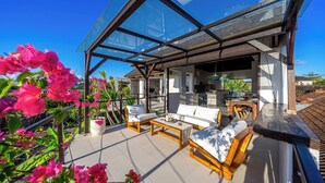 An outdoor patio with wooden furniture and a glass enclosure, surrounded by pink bougainvillea flowers.