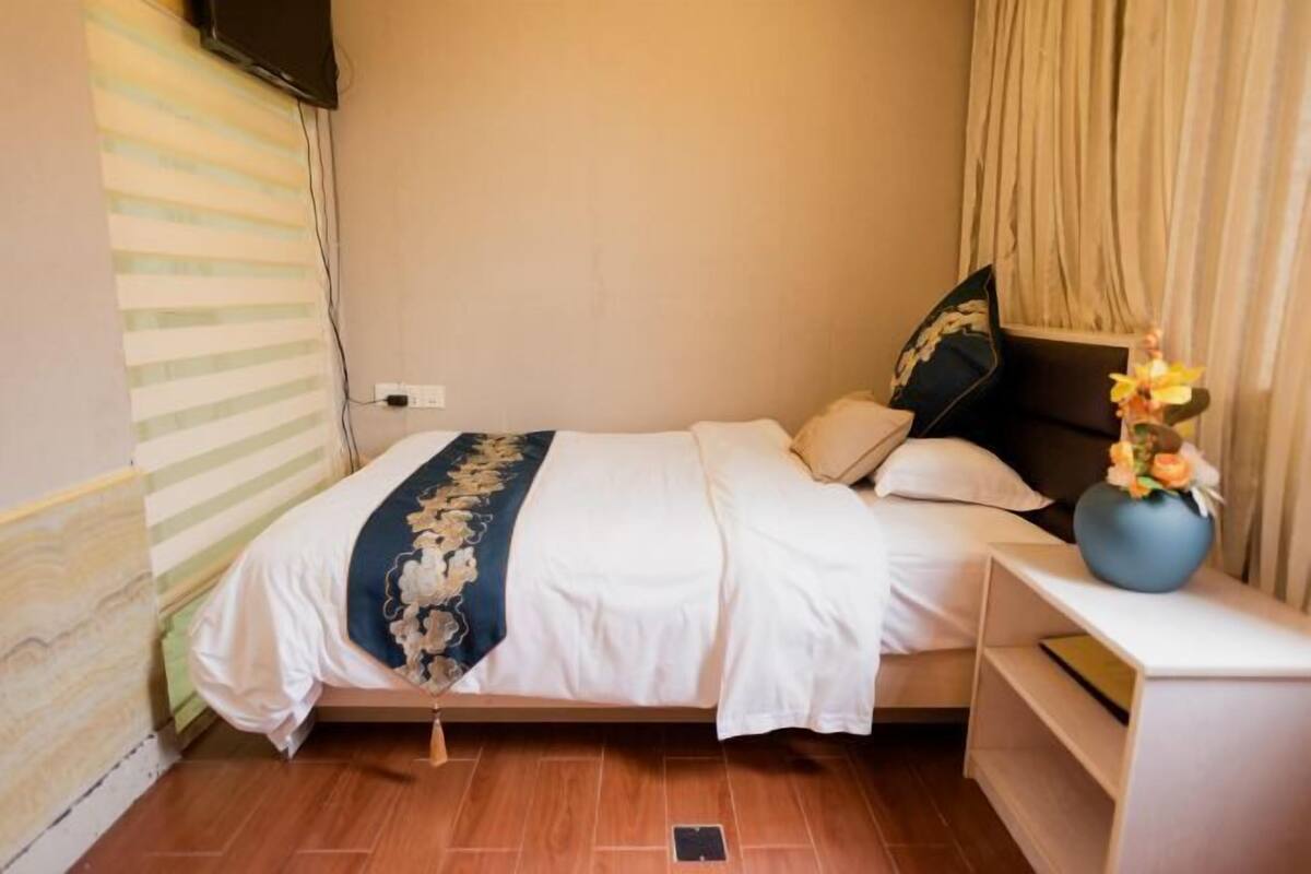 A neatly made bed with a white and blue bedspread, a small bedside table with a vase of flowers, and a wall-mounted TV.