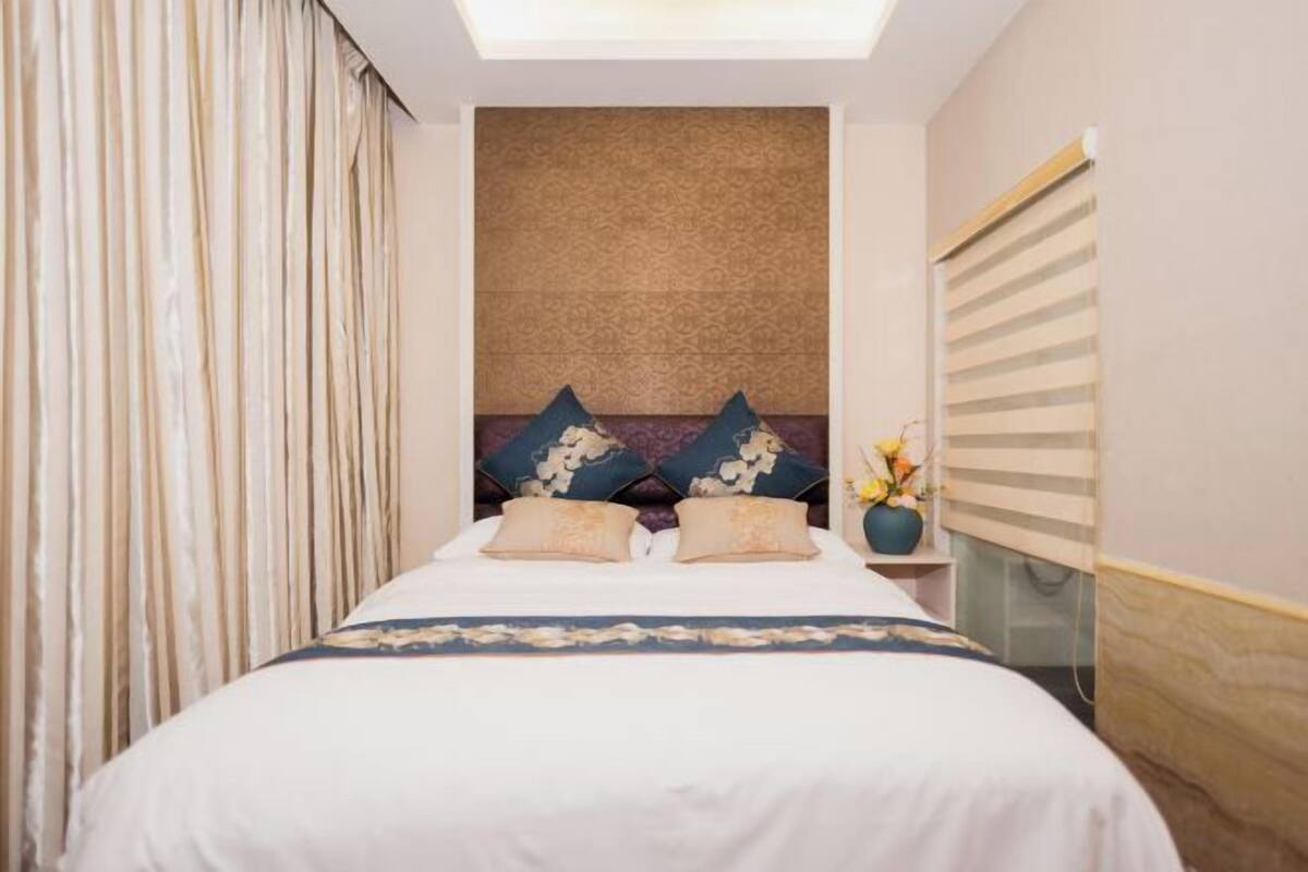 A hotel room with a bed, curtains, a patterned wall, and a nightstand with a vase.