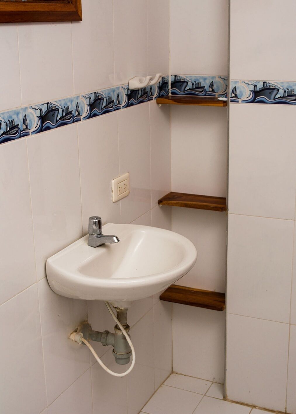 A corner bathroom with a white sink, a faucet, and a pipe. There is a white electrical outlet on the wall.