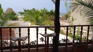 A balcony with wooden railings, a white chair, and a view of a beach with palm trees and a thatched-roof structure. - CASA DE PLAYA FRENTE AL MAR CON JACUZZI  (Cartagena)