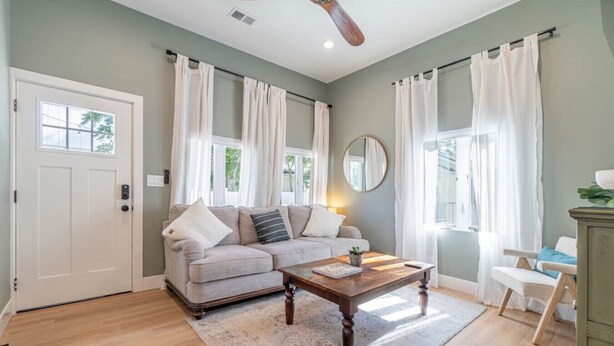 A living room with a sofa, coffee table, and a chair. There is a door, windows with curtains, and a ceiling fan. - Reno Dream Cottage: Delightful 2-Bedroom Sanctuary (Reno)