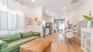 A modern apartment with a green sofa, a brown ottoman, a dining area with a table and chairs, a kitchen with a refrigerator and stove, and a living room with a large window and curtains. - Reno Dream Cottage: Delightful 2-Bedroom Sanctuary (Reno)