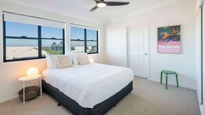 Une chambre à coucher avec un lit, deux fenêtres, un ventilateur de plafond, un luminaire fixé au mur, un tabouret vert et une œuvre d’art encadrée.