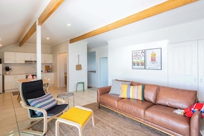 A modern living room with a brown leather sofa, striped cushions, a yellow ottoman, and a dining area with white cabinets and a green stool. - The Hill - Oceanstays (Yamba)