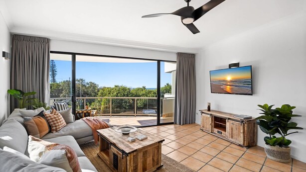 A spacious living room with a large sliding glass door leading to a balcony, a ceiling fan, a flat-screen TV mounted on the wall, and a wooden coffee table. - Pristine 2-Bed Apartment with Pool and Balcony (Sunrise Beach)