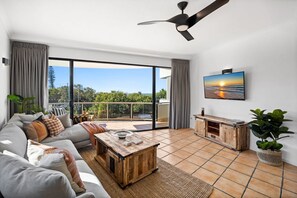 Un salon spacieux doté d’une grande porte-fenêtre coulissante donnant sur un balcon, d’un ventilateur de plafond, d’un téléviseur à écran plat fixé au mur et d’une table basse en bois.