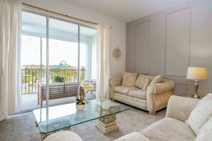 A living room with a glass coffee table, beige sofa, and a view of a balcony with outdoor furniture.