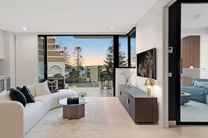 A modern living room with a white sofa, a round coffee table, and a large window offering a view of the outside. - Art-Filled Loft: 50m Beach, King, Parking (Terrigal)