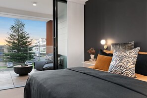 A modern bedroom with a large bed, a nightstand, a lamp, and a view of a tree and buildings outside. - The Loft by Coast Hosting (Terrigal)