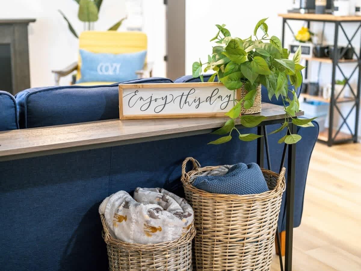 Una sala acogedora con un sofá azul, una cesta de mimbre con un cojín azul, una mesita de mimbre con un letrero que dice "Enjoy this day", y una planta verde.