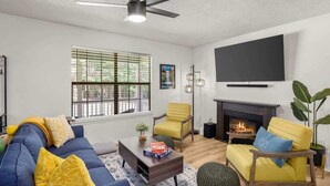 A living room with a sofa, armchairs, a coffee table, a fireplace, and a television mounted on the wall. - Back-40 Retreat with Hot Tub & Bike Garage (Bella Vista)