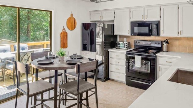 A modern kitchen with white cabinets, a stainless steel refrigerator, a microwave, a stove, and a dining area with a round table and chairs. - Back-40 Retreat with Hot Tub & Bike Garage (Bella Vista)