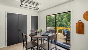 A dining area with a round table, four chairs, and a view of a deck and trees through a large sliding glass door. - Back-40 Retreat with Hot Tub & Bike Garage (Bella Vista)