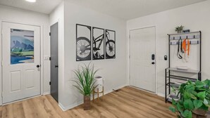 A hallway with wooden flooring, white walls, and three doors. There are three framed pictures on the wall, a plant, and a metal rack with items on it. - Back-40 Retreat with Hot Tub & Bike Garage (Bella Vista)