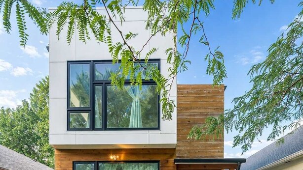 Uma casa moderna de dois andares com grandes janelas, um deck de madeira e um gramado bem cuidado.