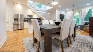 Cozinha moderna com eletrodomésticos de aço inoxidável, área de refeições com mesa de madeira escura e cadeiras brancas, e escada que leva a um andar superior.