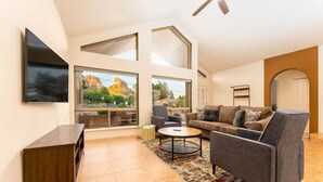 Un salon spacieux avec un grand canapé, une table basse et un téléviseur fixé au mur. De grandes fenêtres offrent une vue sur l’extérieur.