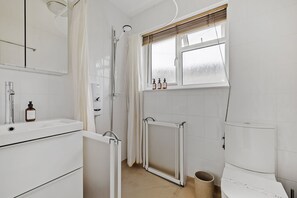 A modern bathroom with a white sink, mirror, and toilet. There is a shower area with a glass door and a window above the toilet.