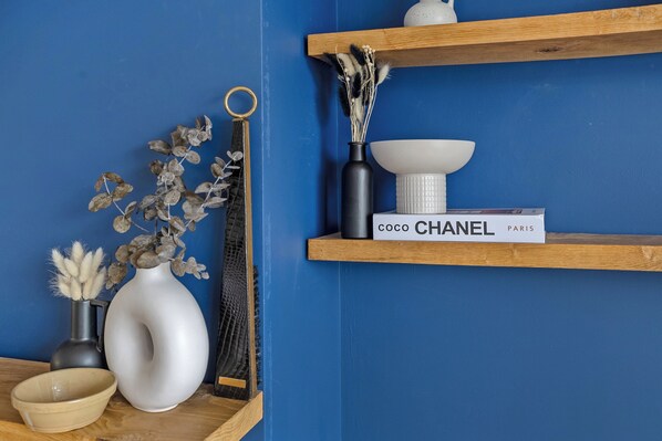 A room with a blue wall, a wooden shelf with a book titled 'coco CHANEL PARIS', a white vase with dried flowers, and a black vase with a black handle.