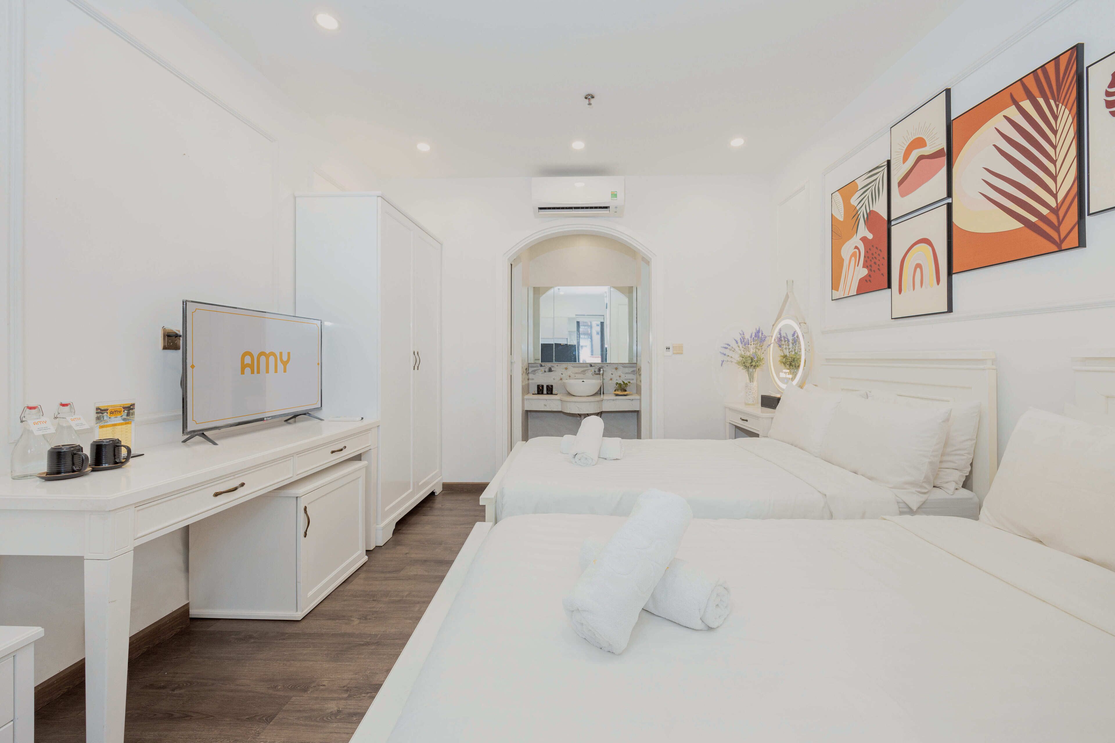 A hotel room with two beds, a desk with a TV, a bathroom with a mirror, and artwork on the wall.