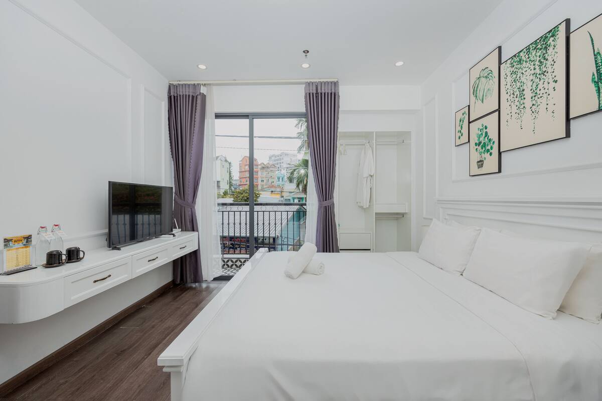 A hotel room with a bed, a TV, a desk, and a balcony with curtains.
