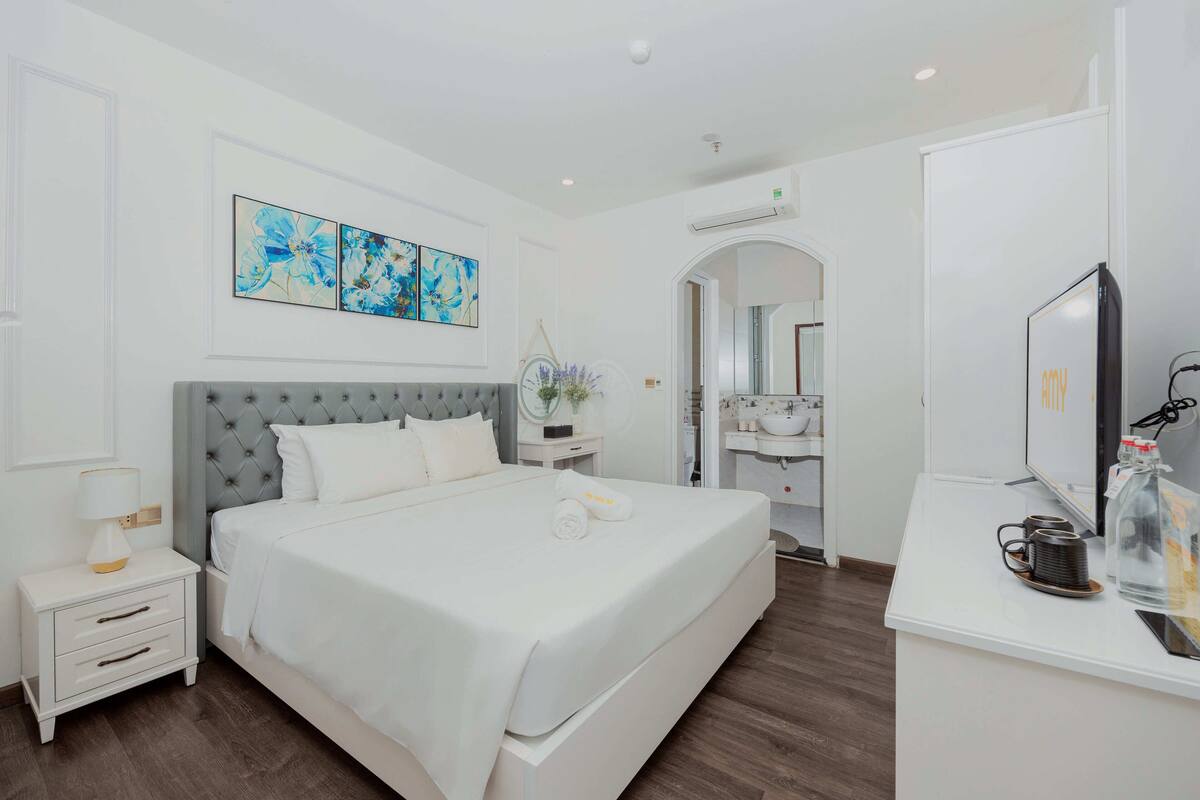 A modern hotel room with a large bed, a bedside table with a lamp, a TV, and a bathroom visible through an archway.