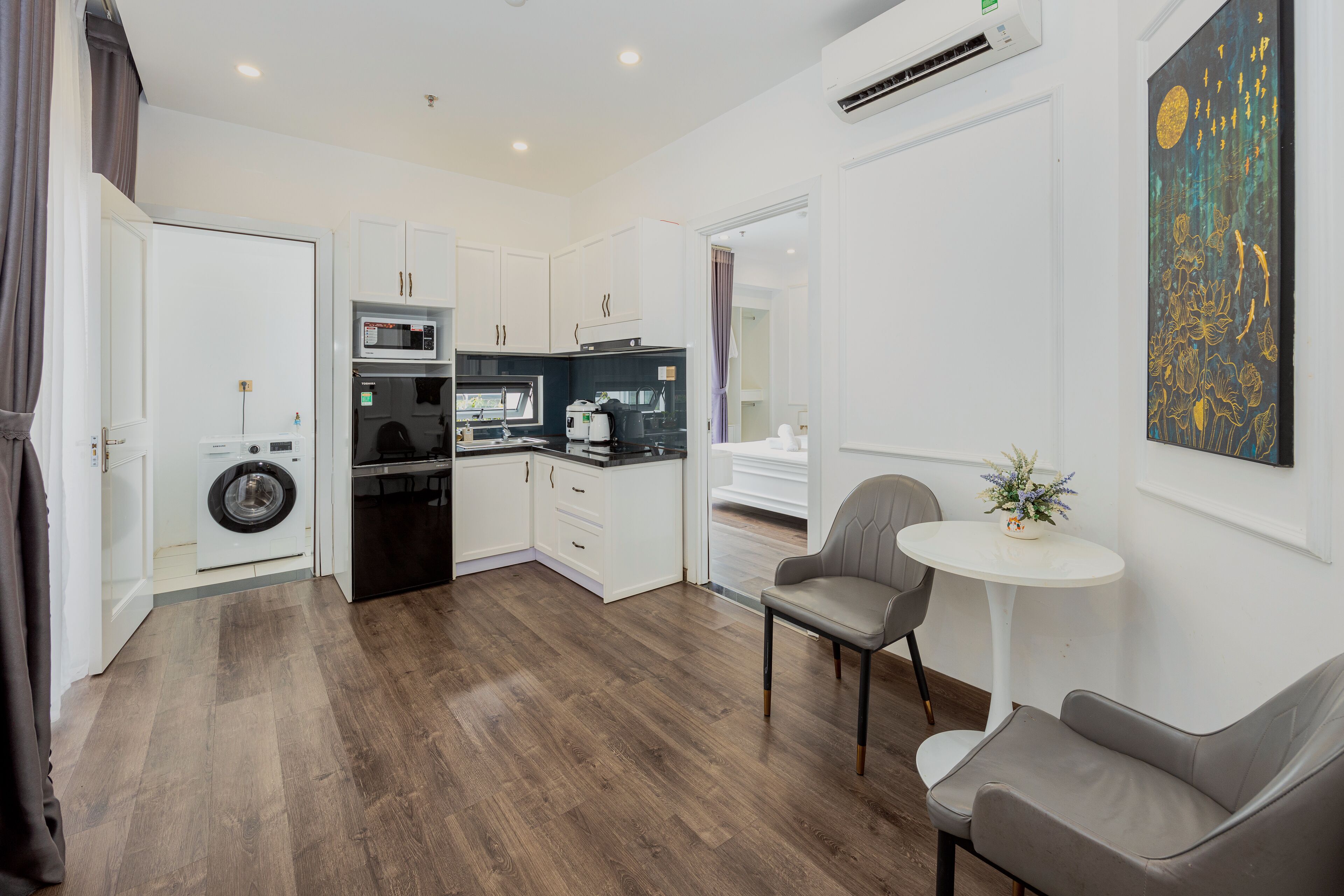 Habitación de hotel pequeña con kitchenette, lavadora, zona de comedor con mesa y sillas, y un cuadro en la pared.