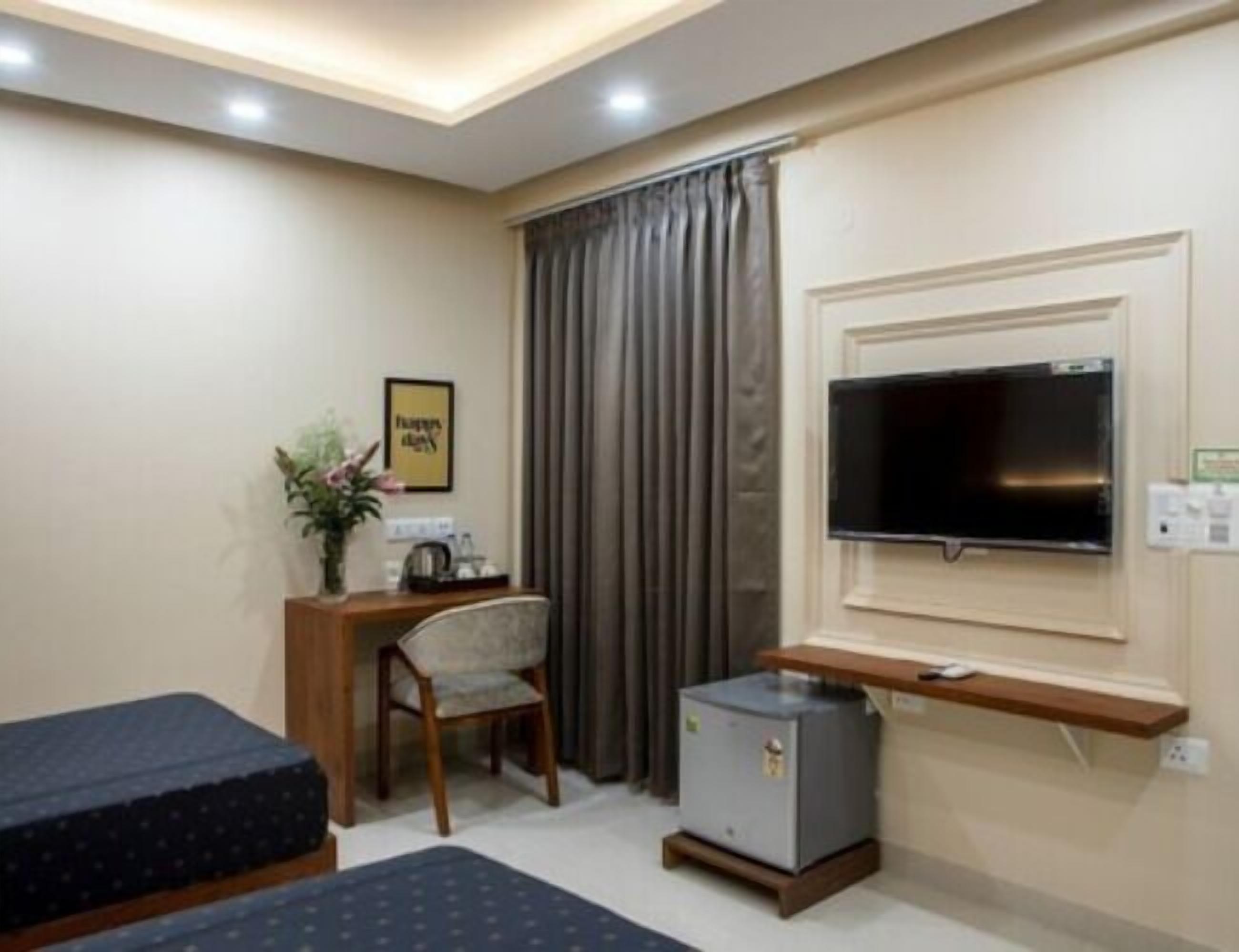 A hotel room with a bed, a desk with a chair, a television mounted on the wall, and a small refrigerator.