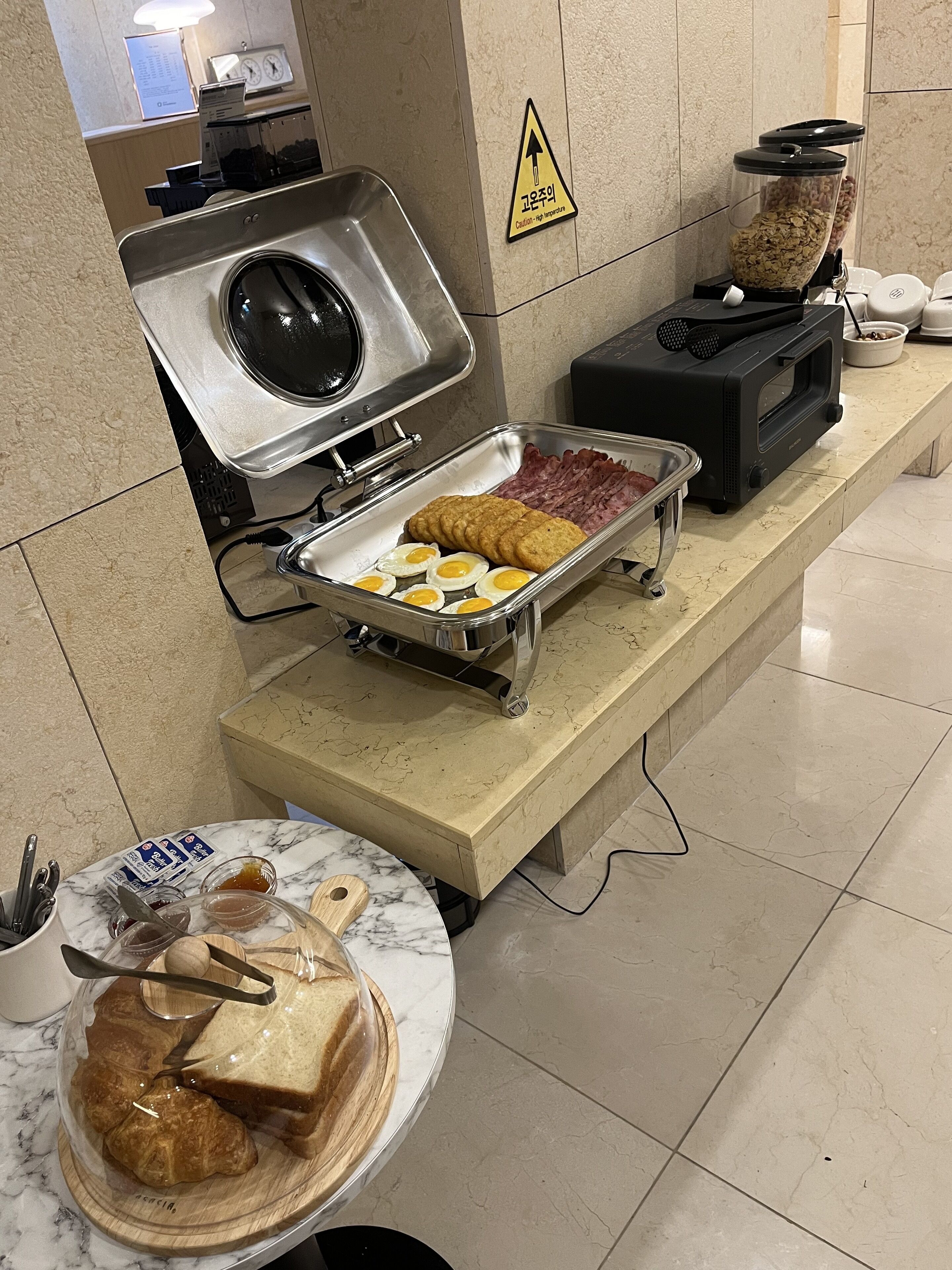 A breakfast buffet with various food items, a coffee machine, and a warning sign on the wall.