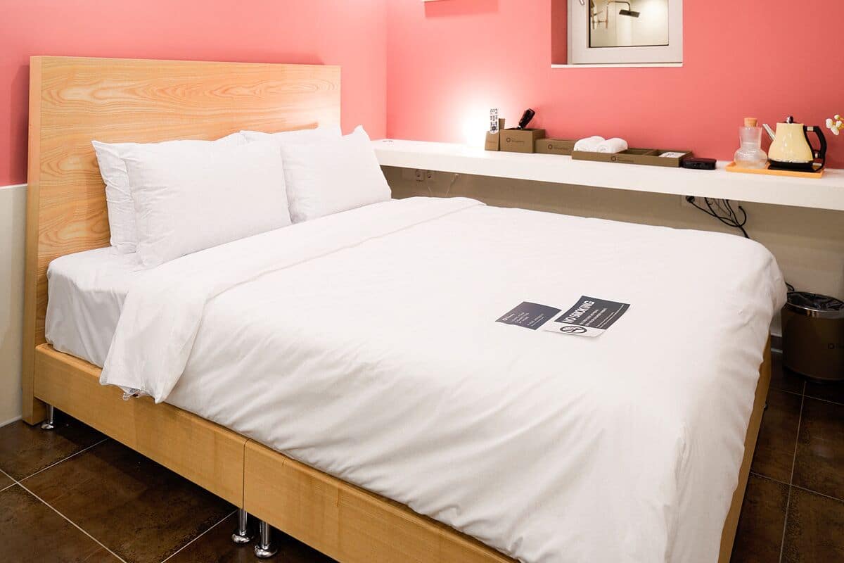 A bedroom with a wooden bed, white bedding, a pink wall, a bathroom mirror, and an air conditioning unit.