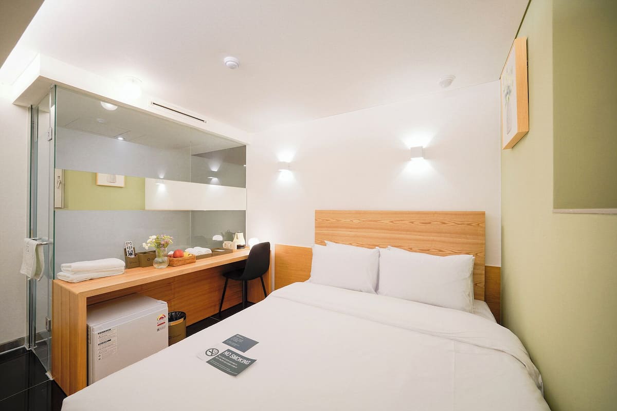 A hotel room with a bed, a desk, a chair, a mirror, and a small refrigerator.